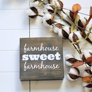 May include: A rustic wooden sign with the text "farmhouse sweet farmhouse" in white paint. The sign is sitting on a white surface with a sprig of cotton bolls.