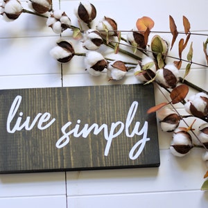 以下が含まれることがあります： 「live simply」の白い文字が書かれた濃い灰色の木製サイン。白い羽目板の背景に置かれています。茶色と白のつぼみと緑と茶色の葉を持つ綿の茎がサインの周りに配置されています。