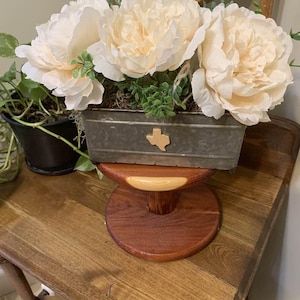 May include: A floral arrangement featuring three large, cream-colored peony blooms in a galvanized metal container. The container has a Texas-shaped accent and sits atop a wooden pedestal. A potted plant is visible to the left.