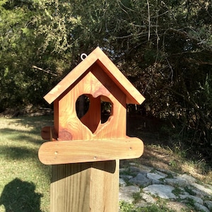 May include: A wooden birdhouse with a heart-shaped opening, mounted on a wooden post. The birdhouse is made of light brown wood and has a pitched roof. The heart-shaped opening is centered on the front of the birdhouse. The birdhouse is designed for outdoor use.
