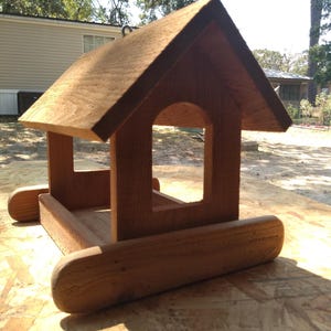 May include: A wooden bird feeder with a gabled roof and rectangular openings. The birdhouse is constructed from light brown wood and features a rounded base. The design resembles a small house, perfect for attracting birds to a garden.