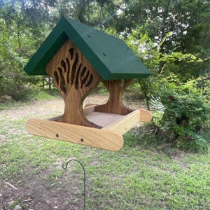 May include: A wooden bird feeder with a green roof and a tree-like design. The feeder is suspended from a hook and has a natural wood finish with a tray for birdseed. The design includes cut-out tree shapes.
