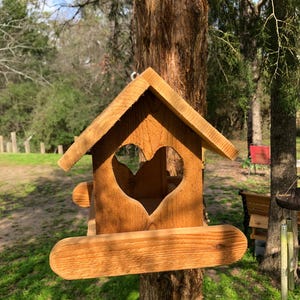 May include: A wooden birdhouse with a heart-shaped cutout and a small perch. The birdhouse is made of light brown wood and is hanging from a tree. The background includes green grass and trees.