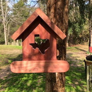 May include: A wooden birdhouse with a Texas-shaped cutout. The birdhouse is made of reddish-brown wood and has a pitched roof. It is mounted on a tree trunk in a natural outdoor setting.