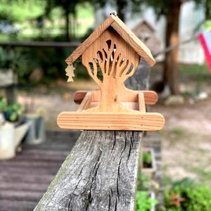 May include: A wooden bird feeder with a tree-shaped cutout design. The birdhouse has a pitched roof and a small decorative cutout hanging from the front. The feeder is made of light-colored wood and is set on a wooden rail.