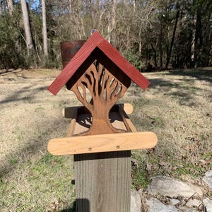 May include: A wooden birdhouse with a red roof and a tree-shaped cutout design. The birdhouse is mounted on a wooden post and has a small perch. The birdhouse is designed to attract birds to a garden or outdoor space.