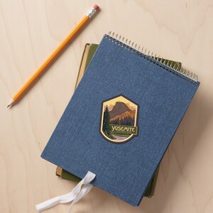Puede incluir: Una libreta azul con espiral y una pegatina vintage del Parque Nacional de Yosemite con una cordillera y un r&iacute;o. La pegatina tiene un fondo dorado y un texto verde que dice "Yosemite National Park".