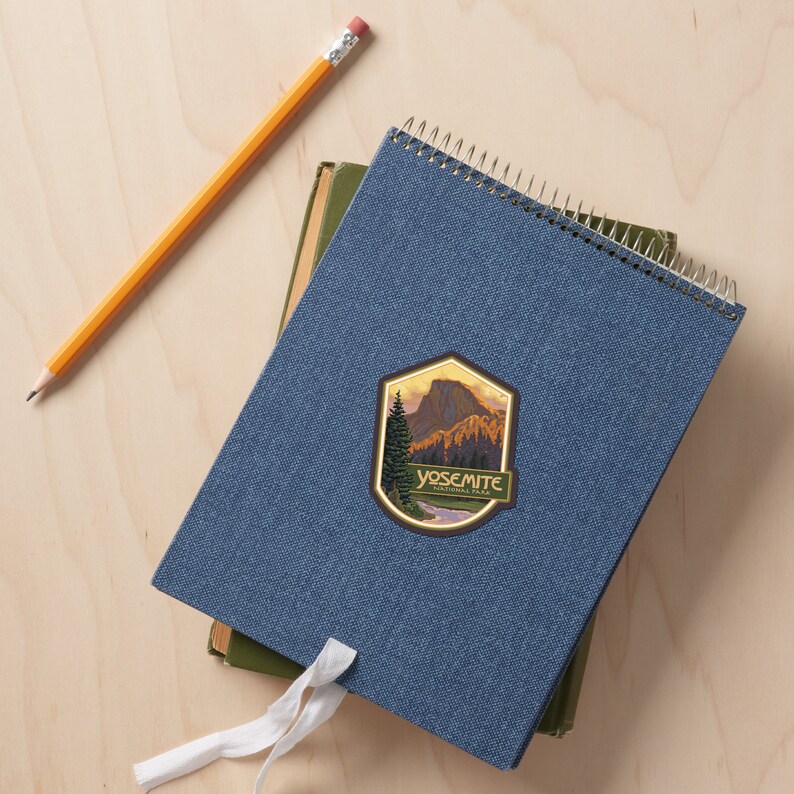Puede incluir: Una libreta azul con espiral y una pegatina vintage del Parque Nacional de Yosemite con una cordillera y un r&iacute;o. La pegatina tiene un fondo dorado y un texto verde que dice "Yosemite National Park".