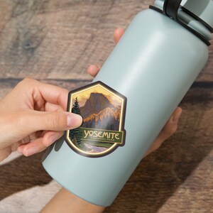 Puede incluir: Una pegatina con una ilustraci&oacute;n vintage de Half Dome en el Parque Nacional de Yosemite. La pegatina es dorada, marr&oacute;n, verde y azul con el texto "Yosemite National Park".