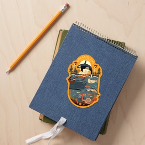 May include: A colorful illustrated sticker featuring an orca whale, a sea otter, a seal, and other marine life. The sticker is on a blue notebook with a white spiral binding. The sticker says "Orcas Island, Washington State".