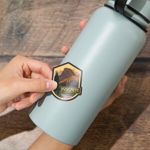 Puede incluir: Una botella de agua azul con una pegatina de una escena de monta&ntilde;a y el texto "Yosemite National Park".