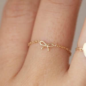 May include: Two gold rings on a finger. One ring has a small bow charm, the other has a heart charm.