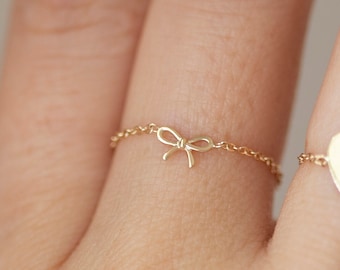 May include: Two gold rings on a finger. One ring has a small bow charm, the other has a heart charm.