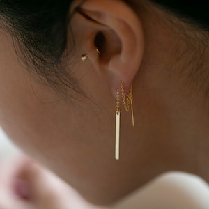 May include: Gold bar earrings with a delicate chain dangling from the bottom. The earrings are worn in the earlobe.