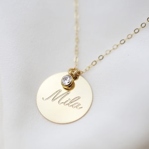 May include: Gold necklace with a round pendant that says "Mila" and a small clear gemstone.