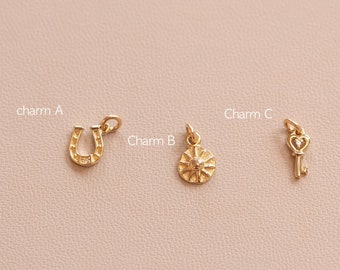 Puede incluir: Tres abalorios de oro: una herradura, un sol y una llave con un agujero en forma de corazón. Los abalorios están sobre un fondo rosa claro.