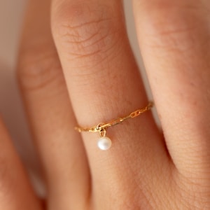 Puede incluir: Un anillo de cadena de oro con una pequeña perla blanca colgando de él. El anillo se lleva en un dedo.