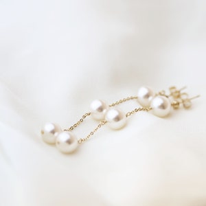 Puede incluir: Un par de pendientes de cadena dorada con perlas blancas. Los pendientes tienen un diseño colgante con varias perlas.