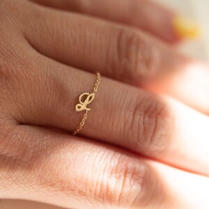 Puede incluir: Un delicado anillo de oro con un dije de letra cursiva 'L'. El anillo se lleva en un dedo.