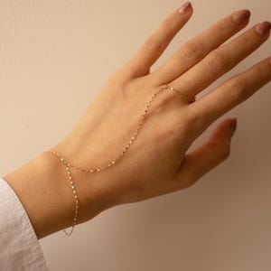 May include: A delicate gold hand chain bracelet. The fine chain connects to a ring and wraps around the back of the hand. The jewellery is displayed against a neutral background, highlighting its elegant design.