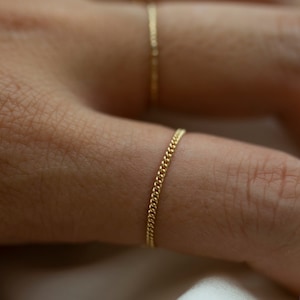 Puede incluir: Un anillo de cadena dorada en un dedo. El anillo está hecho de una cadena delicada con pequeños eslabones redondos.
