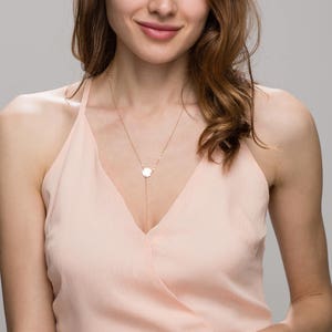 Puede incluir: Una mujer con el pelo largo y castaño lleva un vestido rosa claro sin mangas con un collar de cadena dorada con un pequeño colgante redondo.