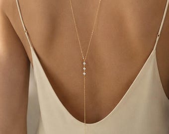 May include: A delicate gold chain necklace worn down the back, featuring a Y-shaped design with three small, clear, round-cut stones set vertically in the center, suspended against a light beige sleeveless garment with thin shoulder straps.