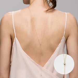 May include: A gold chain necklace with a small ball at the end. The necklace is worn by a person with their back to the camera, wearing a light pink tank top.