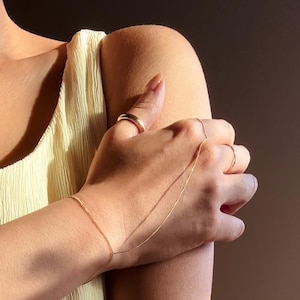May include: A gold-toned hand chain bracelet and rings are worn on a person's hand. The bracelet connects to a ring on the middle finger with a delicate chain. A thick ring is on the index finger, and a thin ring is on the ring finger.