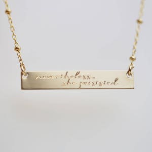 May include: Gold necklace with a bar pendant that reads "nevertheless, she persisted."