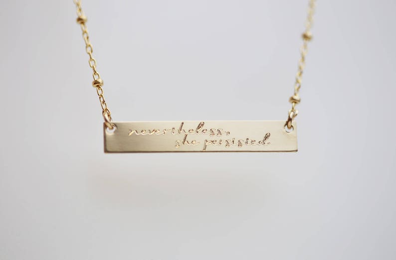 May include: Gold necklace with a bar pendant that reads "nevertheless, she persisted."