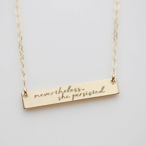May include: Gold necklace with a bar pendant that reads "nevertheless, she persisted."