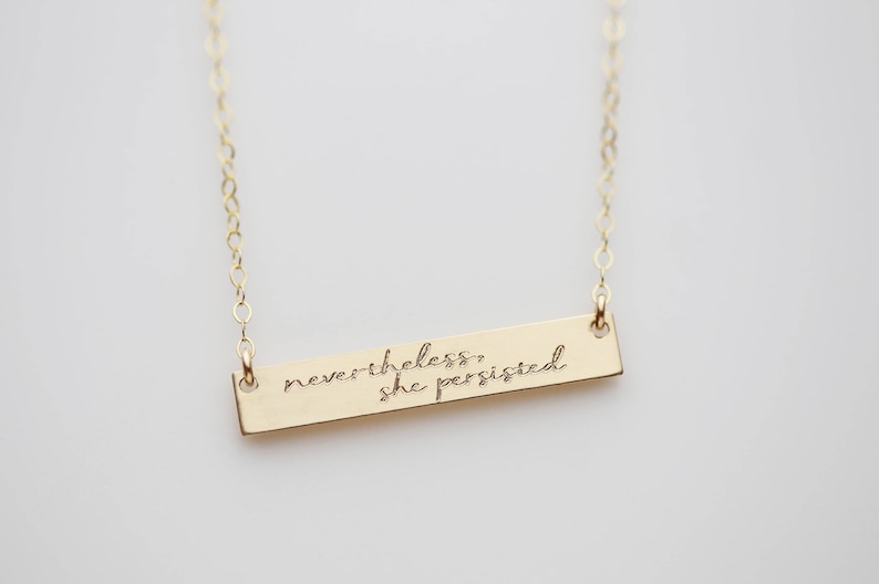May include: Gold necklace with a bar pendant that reads "nevertheless, she persisted."