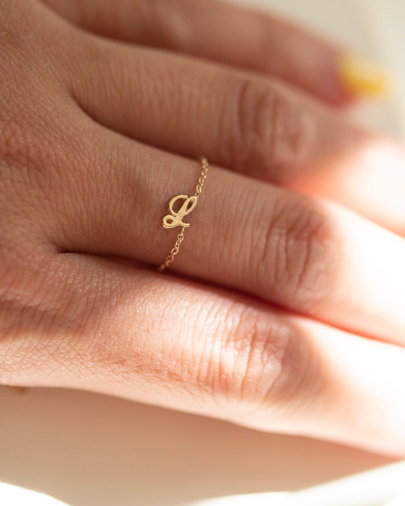 Puede incluir: Un delicado anillo de oro con un dije de letra cursiva 'L'. El anillo se lleva en un dedo.
