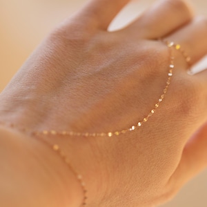 K&ouml;nnte beinhalten: Zarte goldene Handkettenarmband. Das Armband hat eine feine Kette mit kleinen, flachen, kreisf&ouml;rmigen Gliedern. Die Kette verl&auml;uft vom Handgelenk &uuml;ber den Handr&uuml;cken und verbindet sich mit einem Ring. Der Schmuck wird auf einer Hand pr&auml;sentiert.