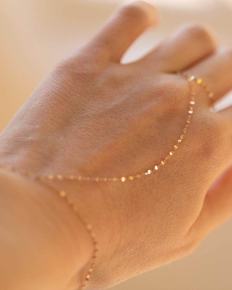 K&ouml;nnte beinhalten: Zarte goldene Handkettenarmband. Das Armband hat eine feine Kette mit kleinen, flachen, kreisf&ouml;rmigen Gliedern. Die Kette verl&auml;uft vom Handgelenk &uuml;ber den Handr&uuml;cken und verbindet sich mit einem Ring. Der Schmuck wird auf einer Hand pr&auml;sentiert.