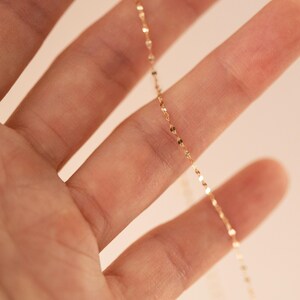 K&ouml;nnte beinhalten: Eine zarte goldene Halskette mit kleinen, flachen, kreisf&ouml;rmigen Anh&auml;ngern. Die Kette wird in der Hand einer Person gehalten, wobei der Fokus auf dem Schmuck liegt. Die Halskette hat einen subtilen Glanz und reflektiert das Licht.