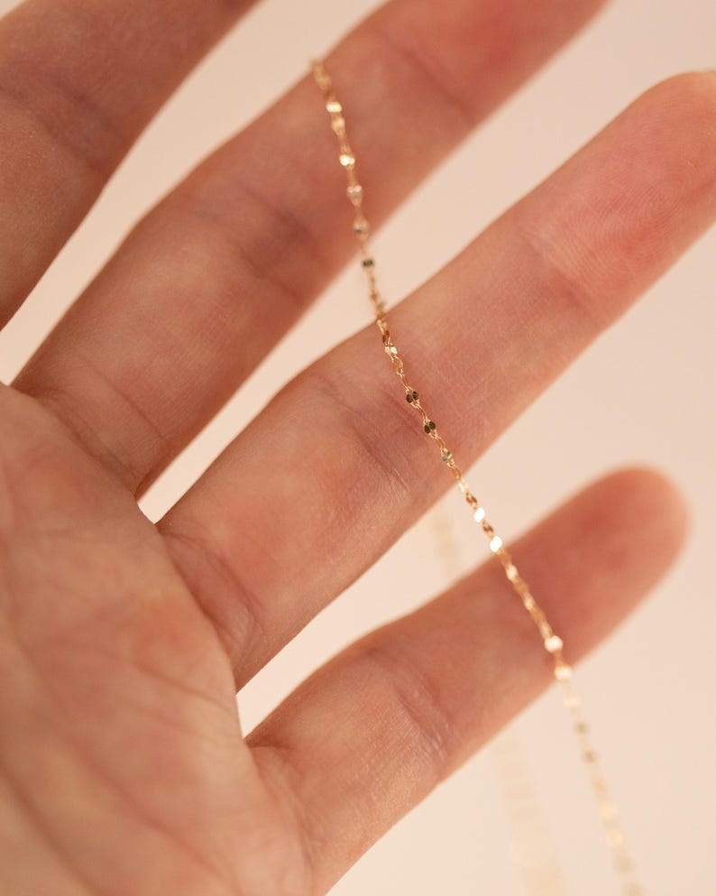 K&ouml;nnte beinhalten: Eine zarte goldene Halskette mit kleinen, flachen, kreisf&ouml;rmigen Anh&auml;ngern. Die Kette wird in der Hand einer Person gehalten, wobei der Fokus auf dem Schmuck liegt. Die Halskette hat einen subtilen Glanz und reflektiert das Licht.