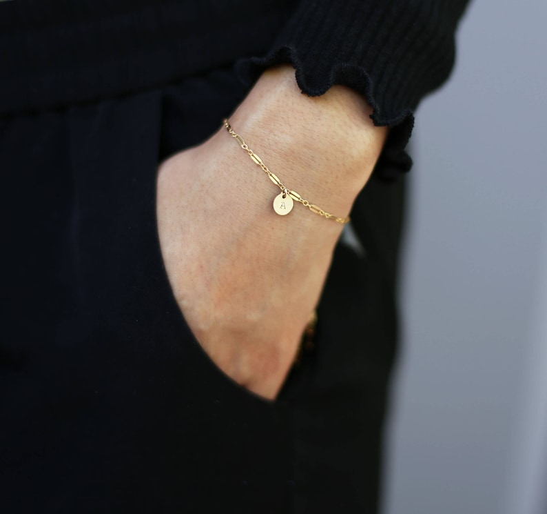 May include: A gold chain bracelet with a small round charm that has the letter 'A' on it.