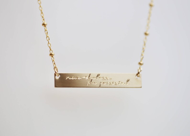 May include: Gold necklace with a bar pendant that reads "nevertheless she persisted".