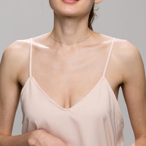 May include: A woman wearing a light pink sleeveless top with a delicate gold chain necklace. The necklace is thin and has a simple design.