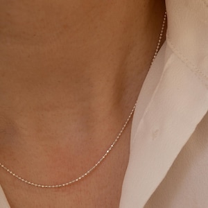 May include: A delicate silver chain necklace with a fine, beaded design. The chain is shown on a person's neck, resting on a white shirt.