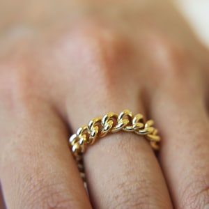 May include: A gold chain ring with a thick, chunky design. The ring is worn on a finger.