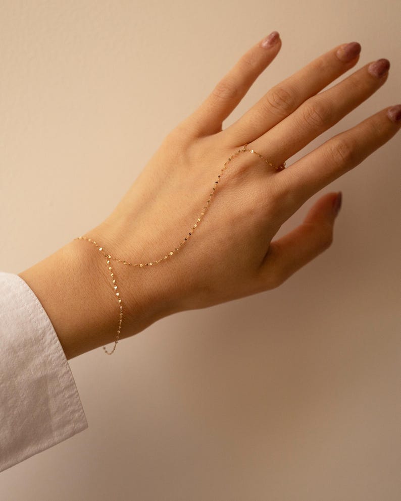 K&ouml;nnte beinhalten: Zarte goldene Handkettenarmband. Die feine Kette verbindet sich mit einem Ring und umschlie&szlig;t den Handr&uuml;cken. Der Schmuck wird vor einem neutralen Hintergrund pr&auml;sentiert, um sein elegantes Design hervorzuheben.