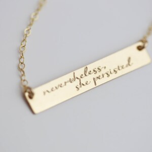 May include: Gold necklace with a bar pendant engraved with the quote "nevertheless, she persisted."