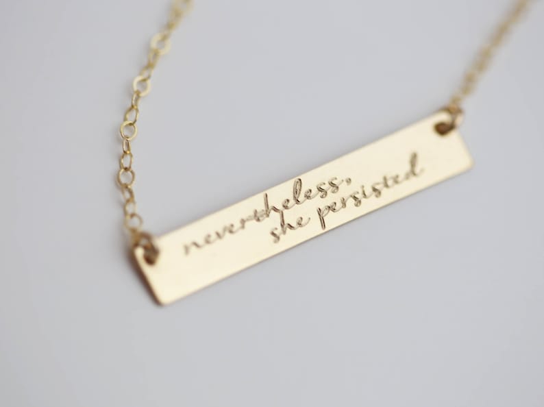 May include: Gold necklace with a bar pendant engraved with the quote "nevertheless, she persisted."
