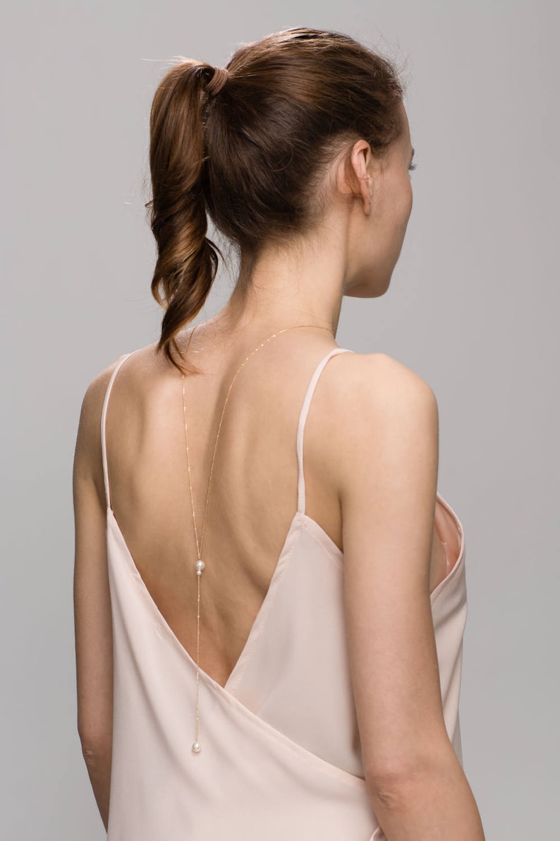May include: A woman wearing a light pink, sleeveless top with a long, delicate gold chain with pearls hanging down her back.