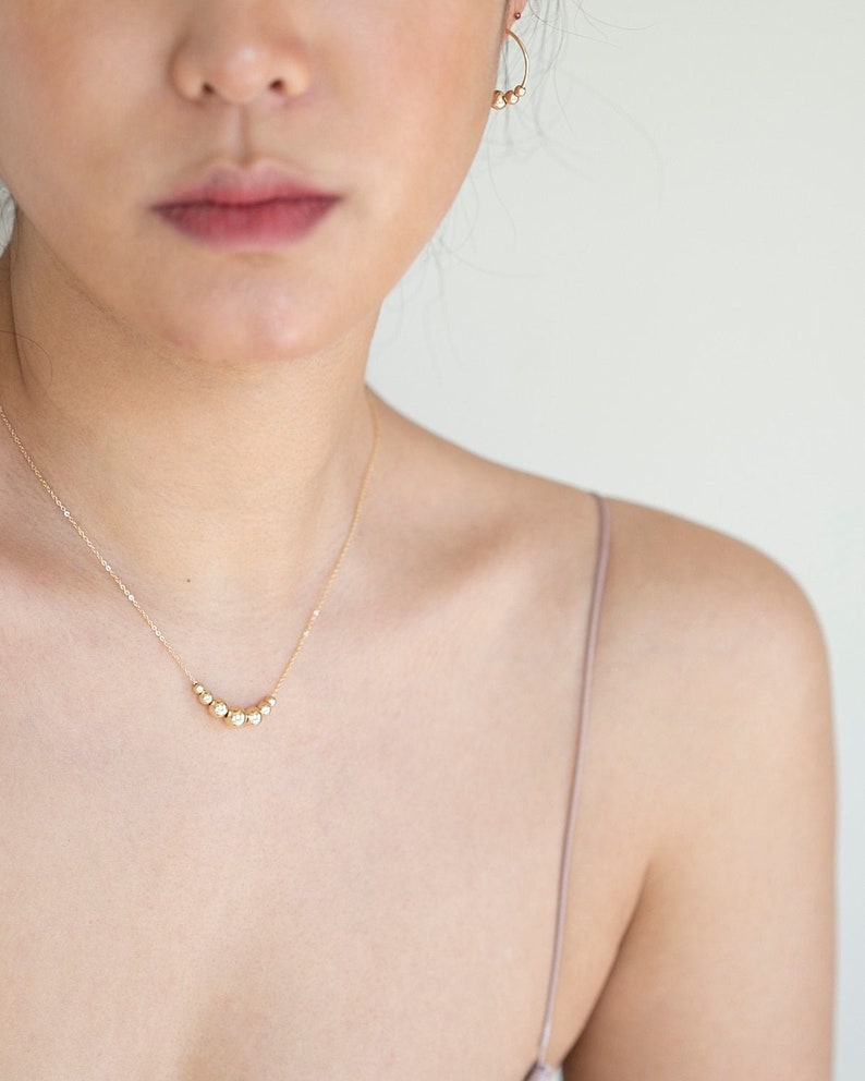 May include: A gold necklace with a delicate chain and a row of small, round beads. The necklace is worn by a person with fair skin.
