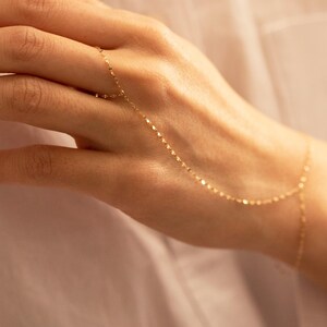 K&ouml;nnte beinhalten: Ein zartes goldenes Handkettenarmband. Das Schmuckst&uuml;ck besteht aus einer feinen Kette, die mit einem Ring verbunden ist und das Handgelenk umschlie&szlig;t. Die Kette besteht aus kleinen, flachen, reflektierenden Gliedern. Das Armband wird an einer Hand gezeigt.
