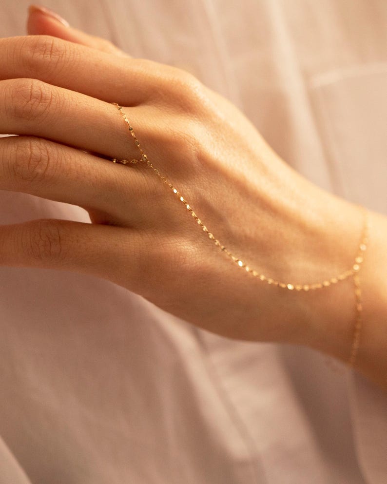 K&ouml;nnte beinhalten: Ein zartes goldenes Handkettenarmband. Das Schmuckst&uuml;ck besteht aus einer feinen Kette, die mit einem Ring verbunden ist und das Handgelenk umschlie&szlig;t. Die Kette besteht aus kleinen, flachen, reflektierenden Gliedern. Das Armband wird an einer Hand gezeigt.